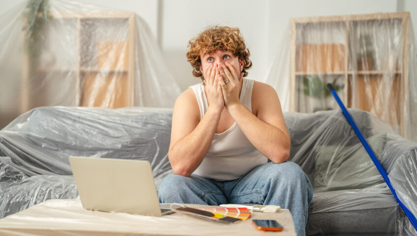 Man looking overwhelmed while planning home renovation with laptop and paint swatches on a covered table