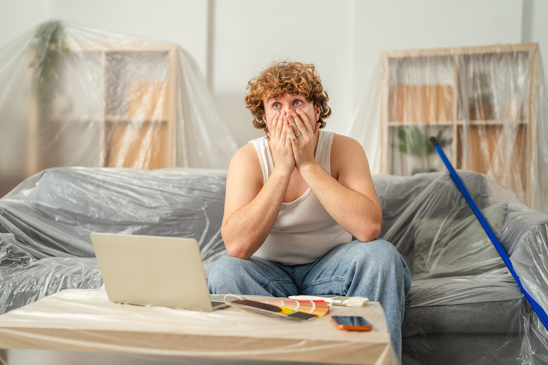 Man looking overwhelmed while planning home renovation with laptop and paint swatches on a covered table